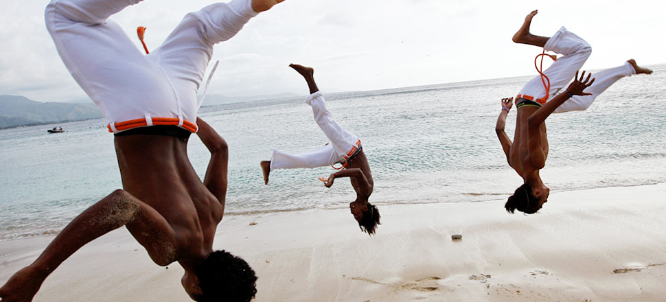 Three shirtless men wearing white pants do flips along the beach