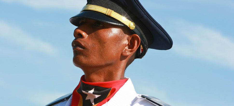 Perfil de soldado com camisa branca, boné preto de oficial e lenço onde se vê uma estrela branca em fundo preto, rodeada de vermelho e amarelo