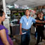 Homem de camisa azul aponta para objeto em uma loja para uma mulher de roupa roxa, com vários outros individíduos observando