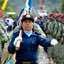 Policial de uniforme azul carrega bandeira à frente de duas fileiras de soldados de uniforme verde