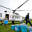 Três caixas azuis em campo aberto, com homem carregando uma quarta caixa tirada de um helicóptero branco da ONU