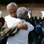 Homem de costas para a câmera, de camisa branca, abraçando outro homem vestido com farda do exército