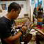Visão lateral de homem pintando, sentado numa cadeira em frente a uma mesa, trabalhando em pintura pequena