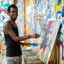Homem sorrindo, de pé, segurando pincel em frente a uma pintura do rosto de um homem, com pinturas coloridas nas paredes do fundo