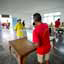 : Visão traseira de homem de camisa vermelha depositando um pedaço de papel em urna eleitoral em cima de uma mesa, observado por mulher de camisa amarela e boné azul
