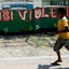 Perfil de menino com camisa amarela e shorte verde, em frente a muro de concreto verde, com letras grandes, brancas e vermelhas: 'Hakribi violencia'